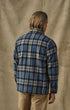 A man viewed from behind wearing a blue, black, and gray plaid flannel shirt and tan pants, standing against a textured beige backdrop.