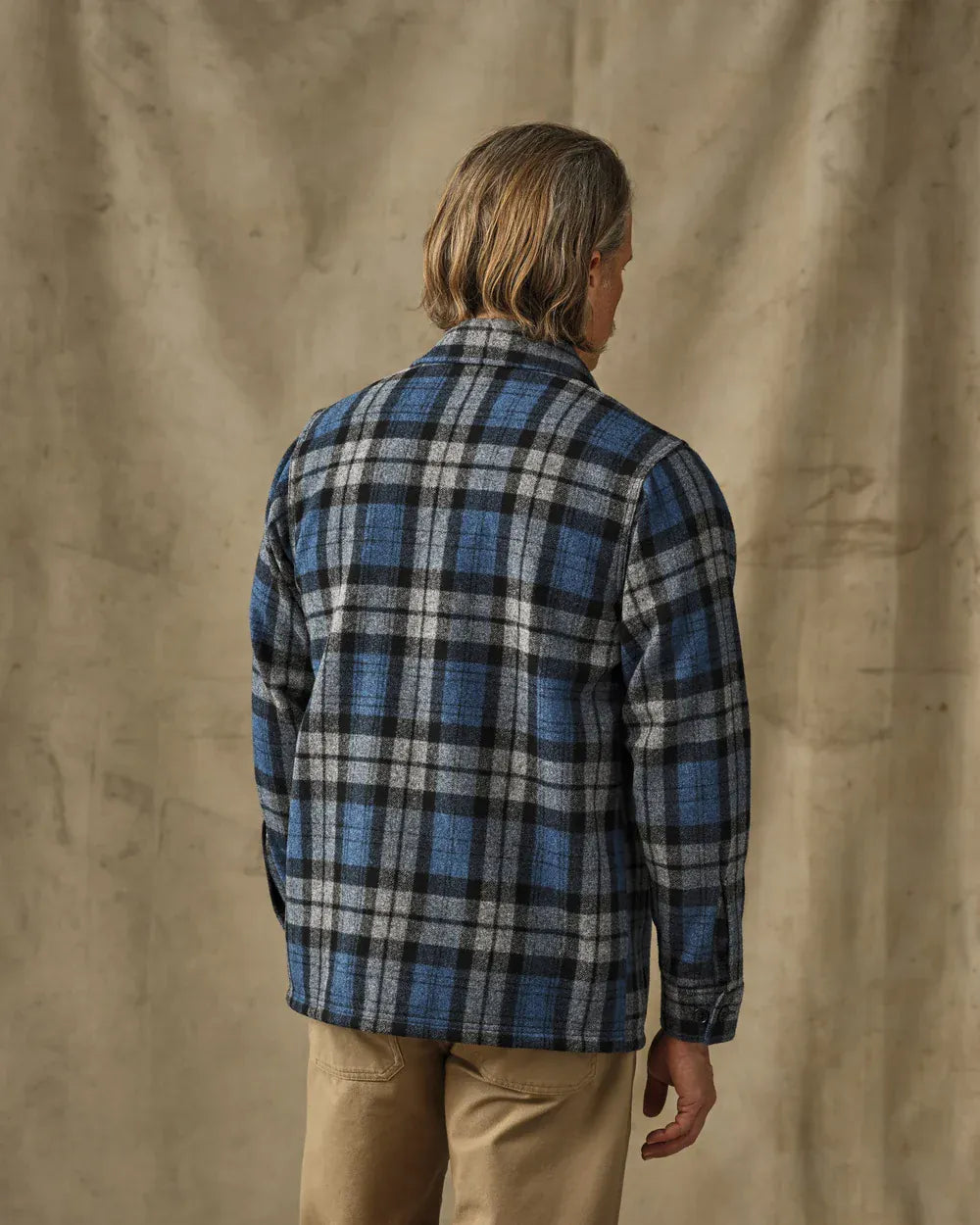 A man viewed from behind wearing a blue, black, and gray plaid flannel shirt and tan pants, standing against a textured beige backdrop.