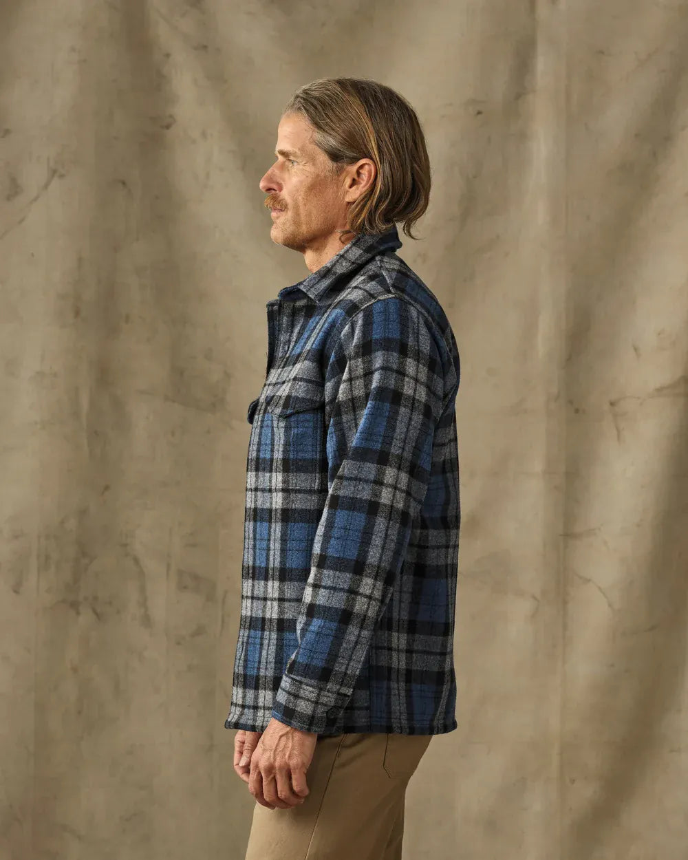 A man in profile wearing a blue and black plaid flannel shirt jacket, paired with tan pants, standing against a textured beige backdrop.