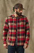 A man wearing a red, black, and beige plaid flannel button-up shirt, a brown trucker hat, and dark blue jeans, standing against a textured brown backdrop.