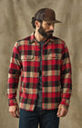 A man wearing a red, black, and beige plaid flannel button-up shirt, a brown trucker hat, and dark blue jeans, standing against a textured brown backdrop.
