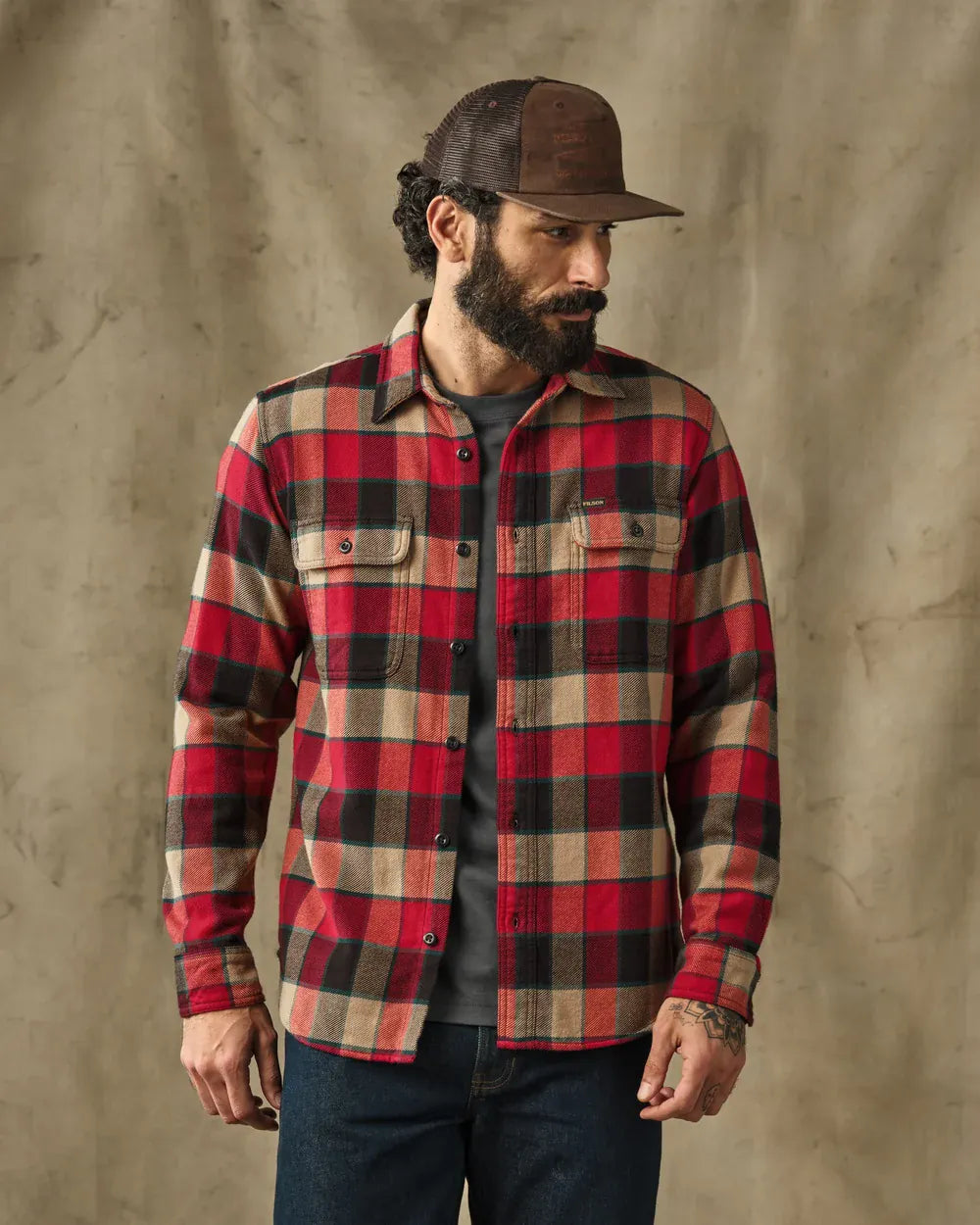 A man wearing a red, black, and beige plaid flannel button-up shirt, a brown trucker hat, and dark blue jeans, standing against a textured brown backdrop.