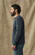 A man with a beard and curly hair stands in profile wearing a dark slate blue long-sleeve henley shirt and blue jeans, set against a textured beige fabric backdrop.