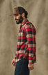 A man in profile wearing a red, black, and beige plaid flannel shirt, a brown trucker hat, and dark blue jeans, standing against a textured beige backdrop.