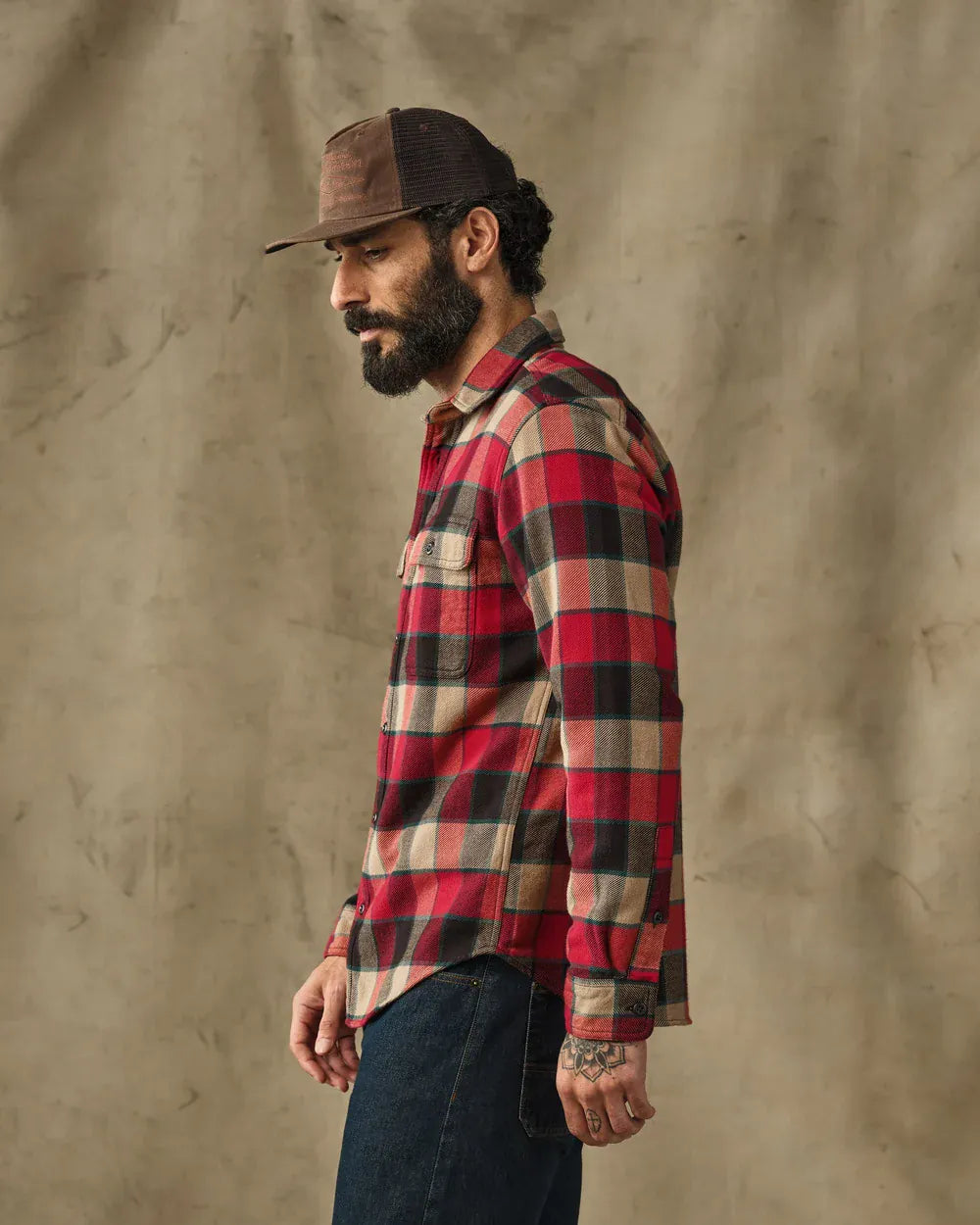 A man in profile wearing a red, black, and beige plaid flannel shirt, a brown trucker hat, and dark blue jeans, standing against a textured beige backdrop.