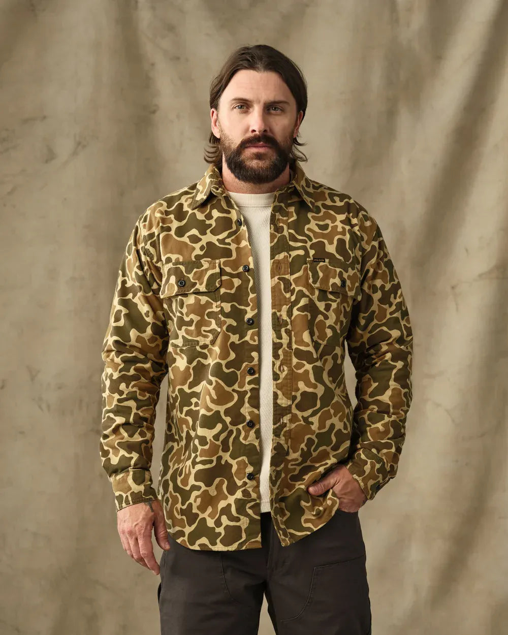 A man with a beard modeling a camouflage patterned button-up shirt over a white t-shirt and dark pants, standing against a textured beige backdrop.