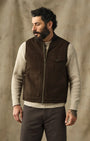 A bearded man wearing a dark brown utility zip-front vest over a cream ribbed sweater and dark pants, standing against a textured beige backdrop.