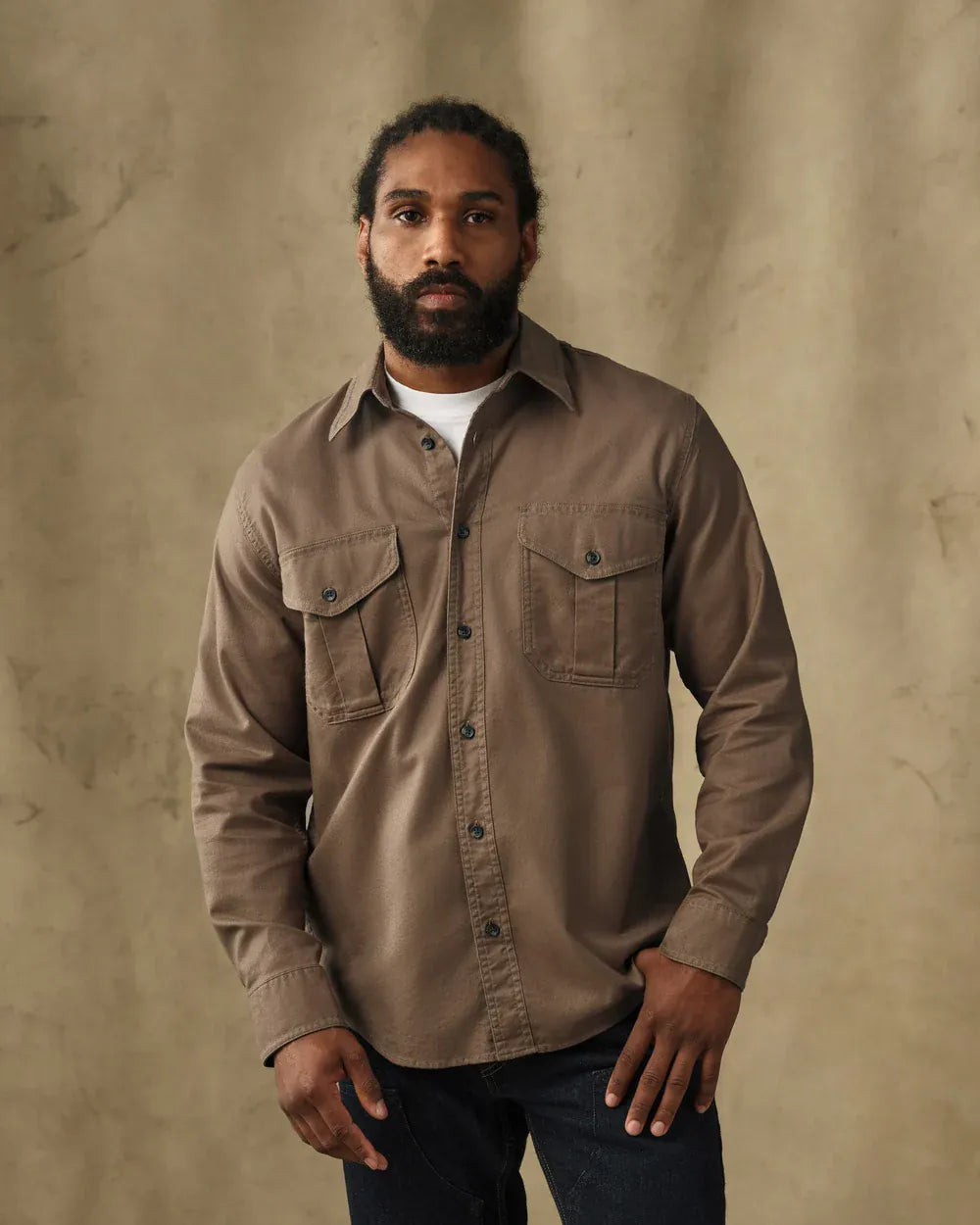 A man wearing a brown long-sleeve button-up shirt with two chest pockets, styled over a white t-shirt and dark jeans, standing against a textured beige backdrop.