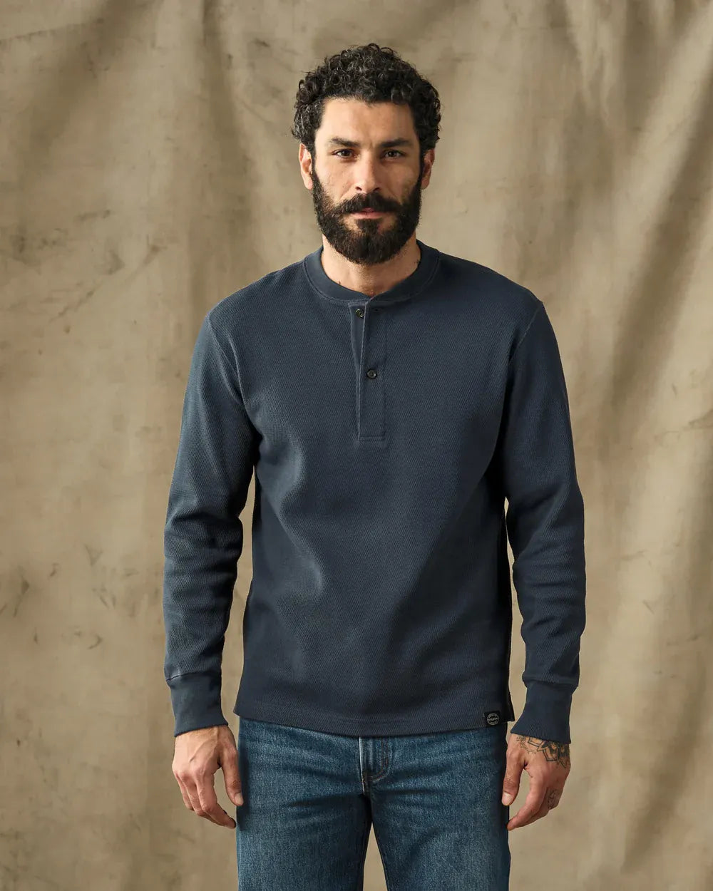 A man with a beard wearing a dark slate blue long-sleeve henley shirt and blue jeans, standing against a textured beige backdrop.