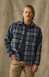 A man wearing a blue, gray, and black plaid button-up flannel shirt over a dark t-shirt and khaki pants, standing against a textured beige backdrop.