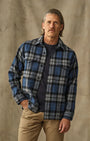 A man wearing a blue, gray, and black plaid button-up flannel shirt over a dark t-shirt and khaki pants, standing against a textured beige backdrop.
