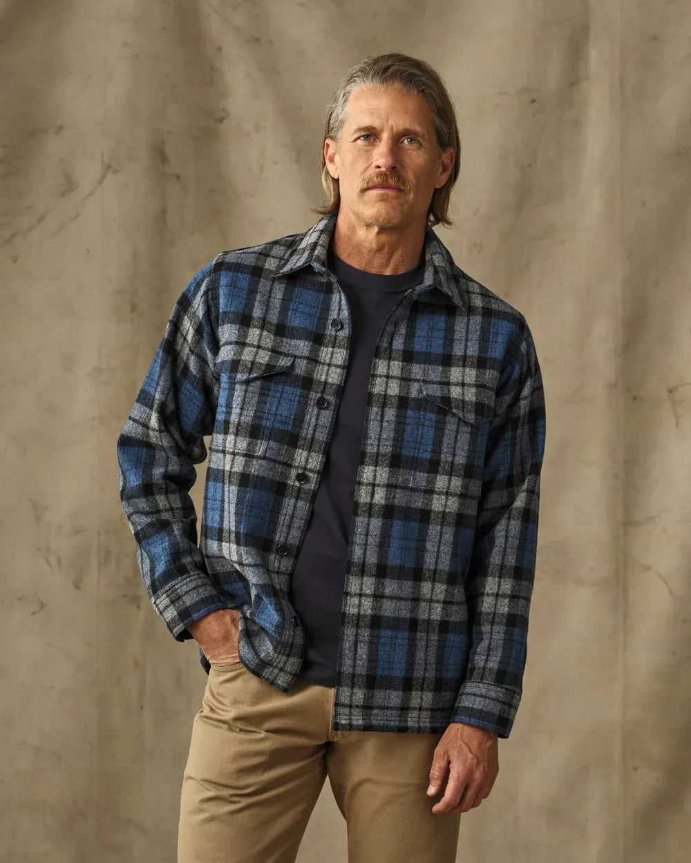 A man wearing a blue, gray, and black plaid button-up flannel shirt over a dark t-shirt and khaki pants, standing against a textured beige backdrop.