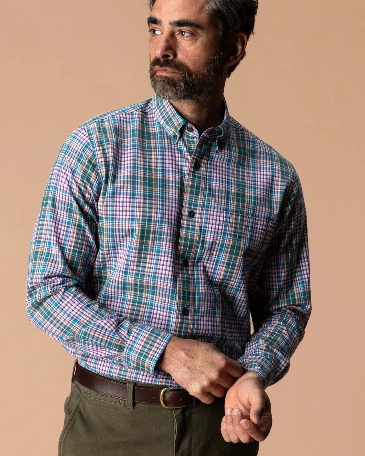 A man wearing a long-sleeve button-down plaid shirt in green, blue, and purple tones, paired with olive green trousers, against a solid beige background.