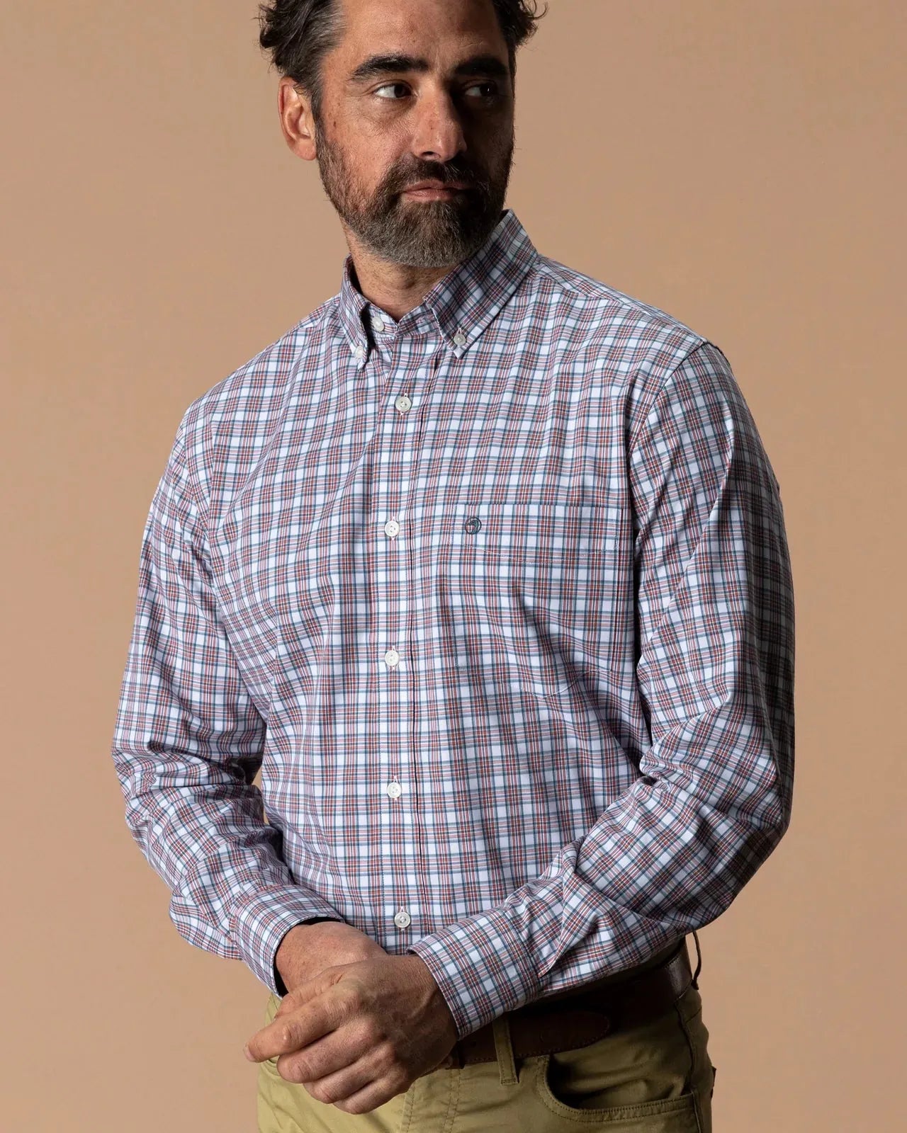 A man wearing a long-sleeve button-down shirt with a blue, red, and white plaid pattern, paired with khaki pants, set against a solid beige background.