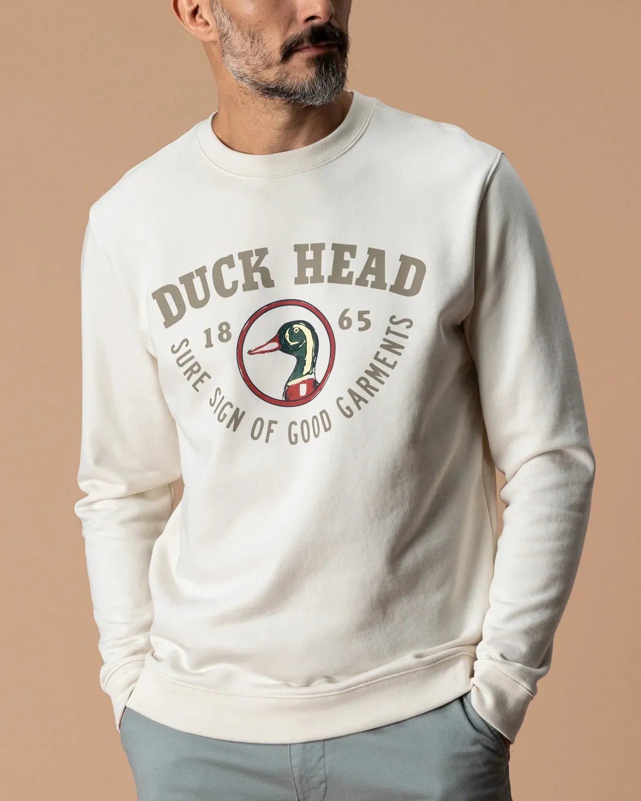 A man wearing a cream-colored long-sleeve crewneck sweatshirt with a vintage-style 'Duck Head' graphic on the chest, paired with light gray pants, set against a solid beige background.