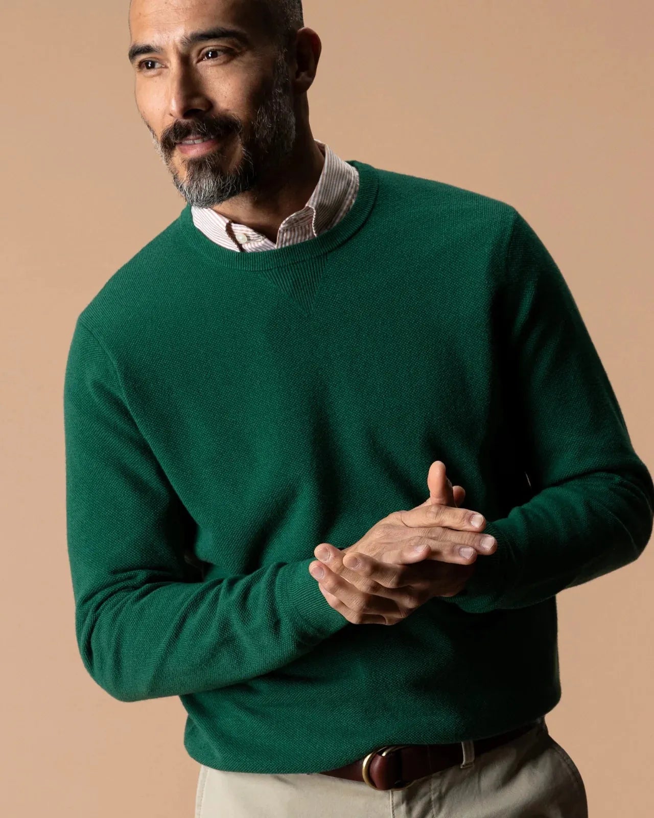 A man wearing a dark green crewneck sweater over a light-colored collared shirt, paired with beige trousers, posed against a solid warm beige background.