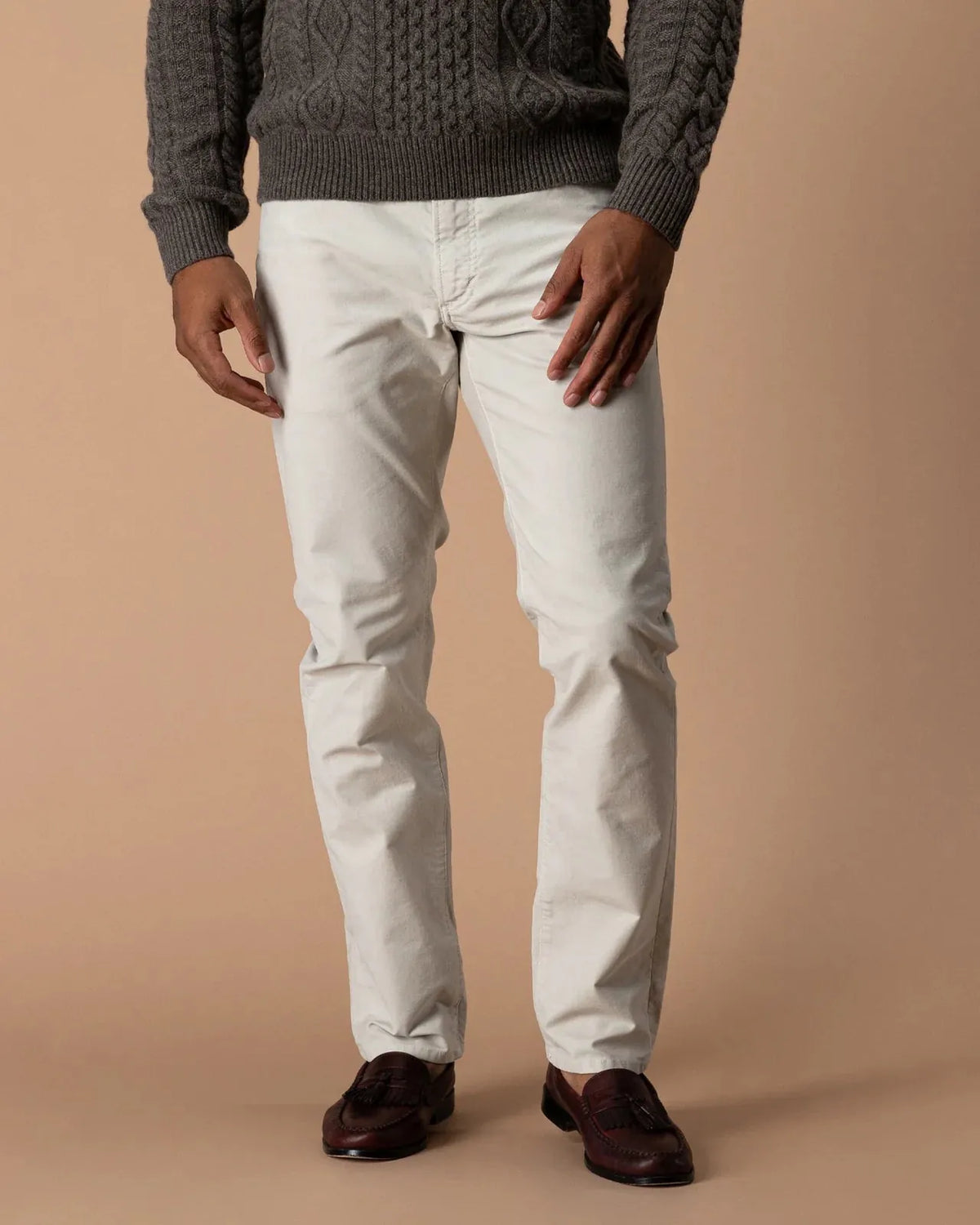 A person wearing off-white slim-fit trousers, a dark gray cable-knit sweater, and dark brown leather loafers, standing against a solid beige studio background.
