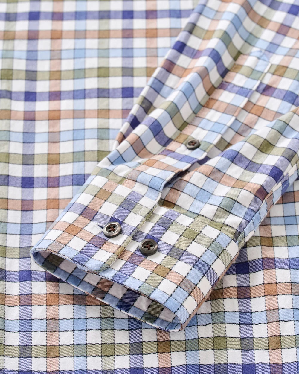 Close-up of a plaid button-up shirt sleeve with a multicolor check pattern in blue, purple, green, beige, and white, featuring dark buttons.