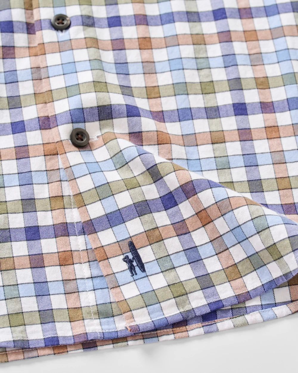 Close-up of a plaid button-up shirt with a multicolor check pattern in blue, purple, green, and peach on a white base, featuring a small embroidered polo player logo near the cuff.
