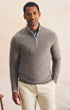 A man wearing a light gray quarter-zip knit sweater over a collared shirt and cream trousers, standing against a warm beige textured wall.
