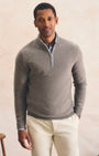 A man wearing a light gray quarter-zip knit sweater over a collared shirt and cream trousers, standing against a warm beige textured wall.