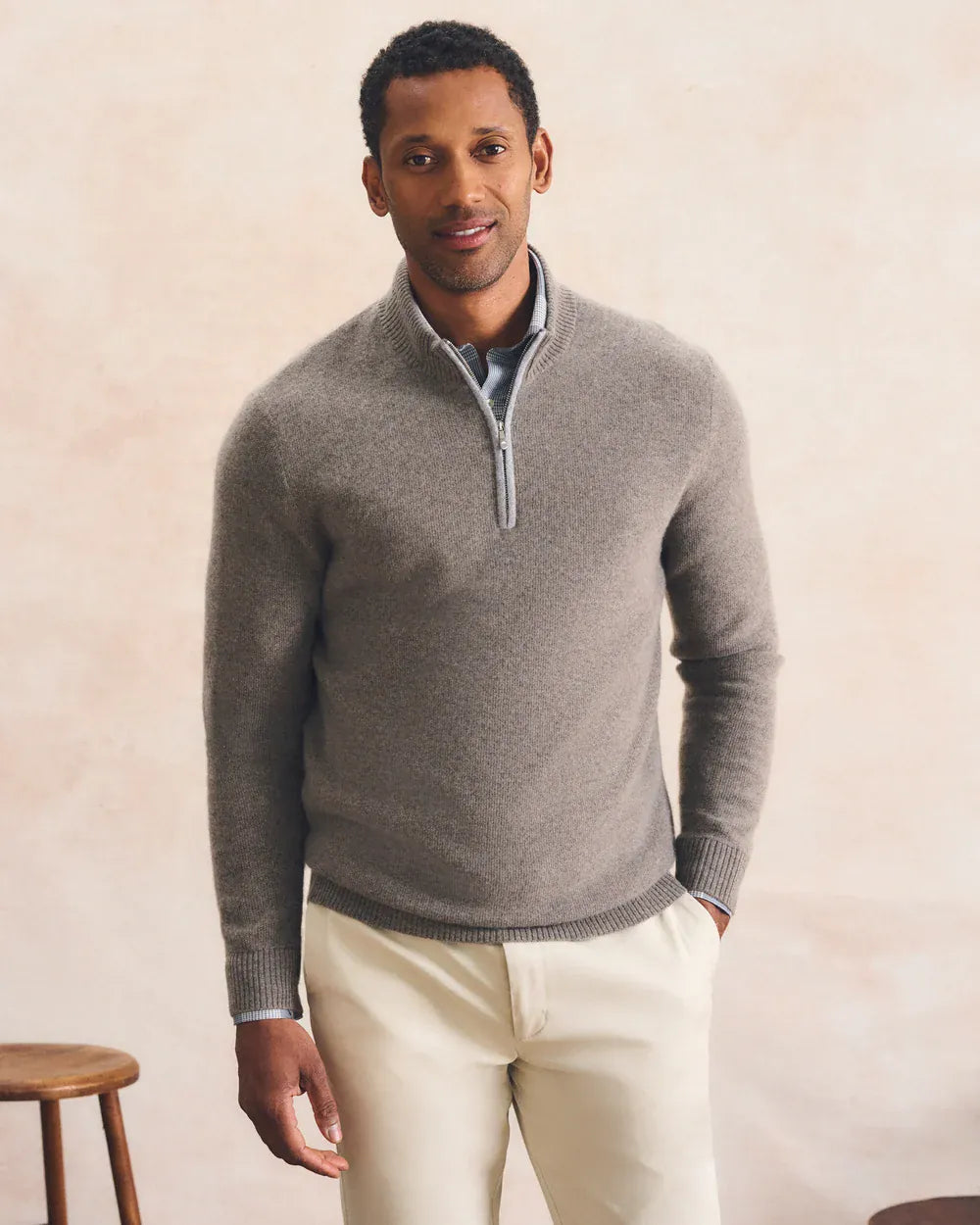 A man wearing a light gray quarter-zip knit sweater over a collared shirt and cream trousers, standing against a warm beige textured wall.