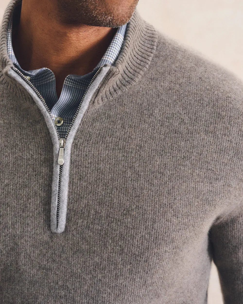 Close-up of a man wearing a light gray ribbed knit quarter-zip sweater over a blue and white checkered collared shirt.