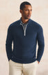 A man wearing a navy blue quarter-zip knit sweater over a patterned collared shirt and cream pants, standing against a textured beige background.