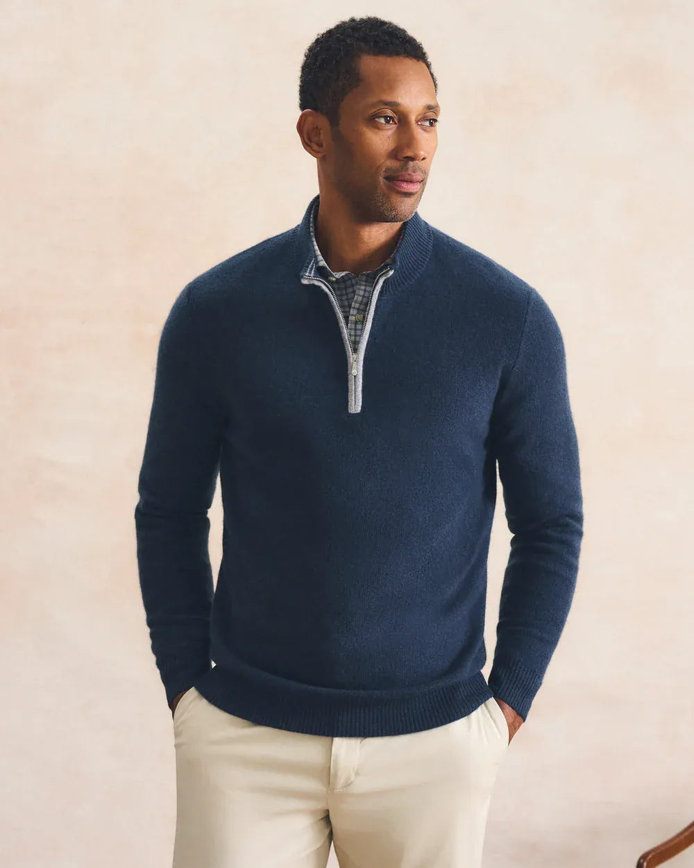 A man wearing a navy blue quarter-zip knit sweater over a patterned collared shirt and cream pants, standing against a textured beige background.