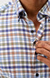 Close-up of a man wearing a long-sleeve button-down shirt with a multicolor plaid pattern in blue, green, brown, and white. He is adjusting the collar with one hand.