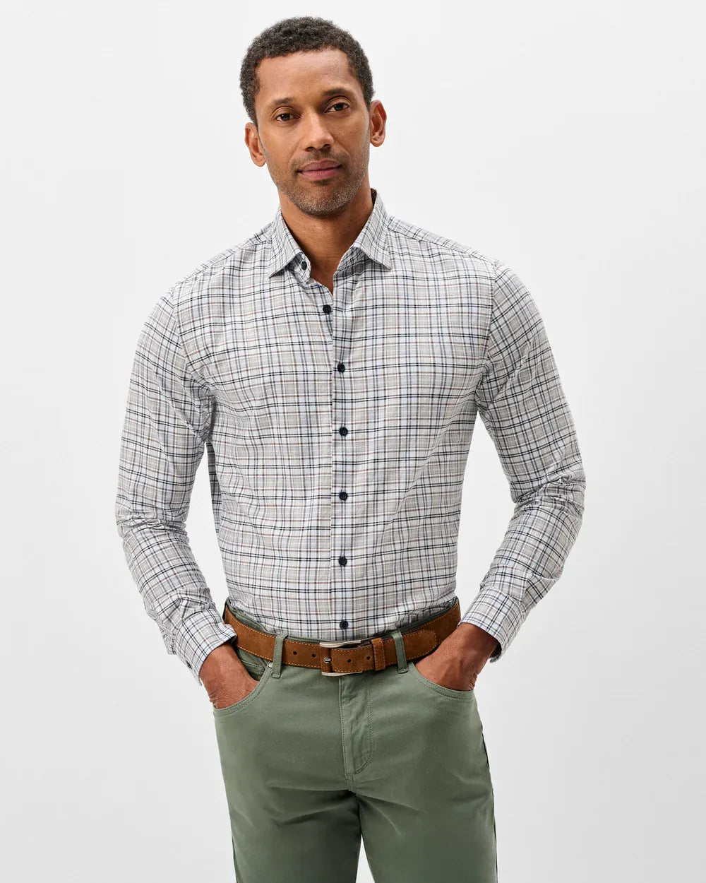 A man wearing a long-sleeve button-down shirt with a gray, white, and black plaid pattern, styled with olive green trousers and a brown leather belt, against a plain light gray background.