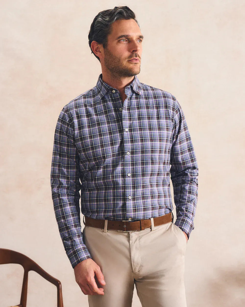 A man wearing a blue and gray plaid button-down shirt, beige trousers, and a brown belt, standing against a textured beige wall.