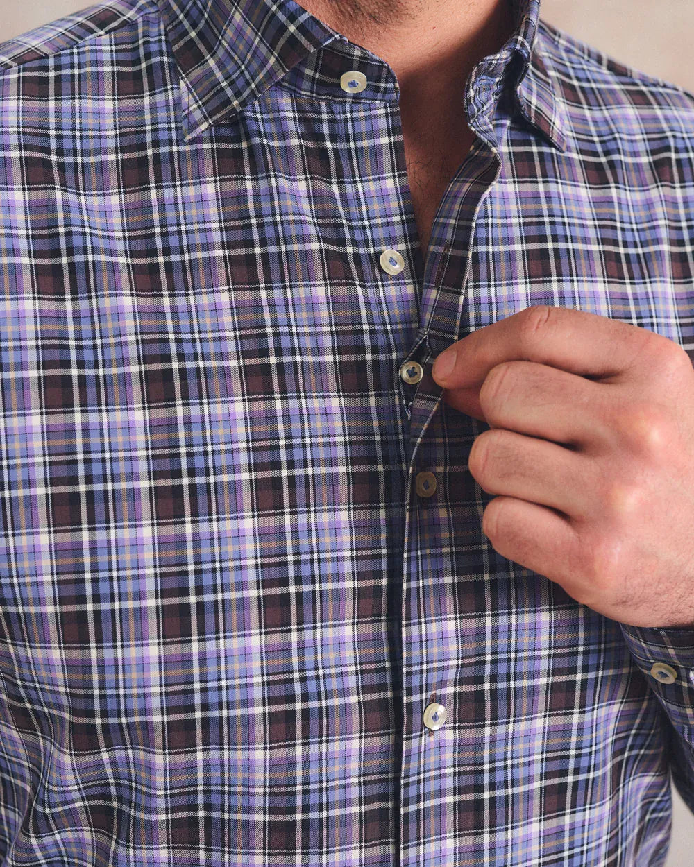A close-up of a man wearing a plaid button-down shirt, adjusting the collar and buttoning the front. The shirt features a purple, black, and white plaid pattern.