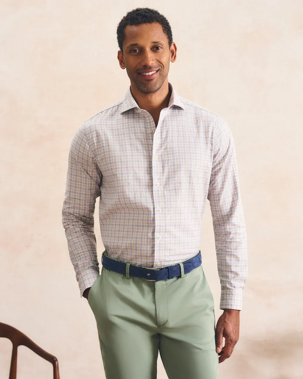 A man wearing a long-sleeve button-down dress shirt with a fine blue and beige check pattern, paired with sage green trousers and a dark blue belt, standing against a textured beige background.