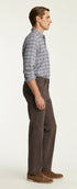 A man shown in profile wearing a blue and gray plaid button-down shirt, dark brown trousers, and brown suede shoes, standing against a plain off-white studio background.