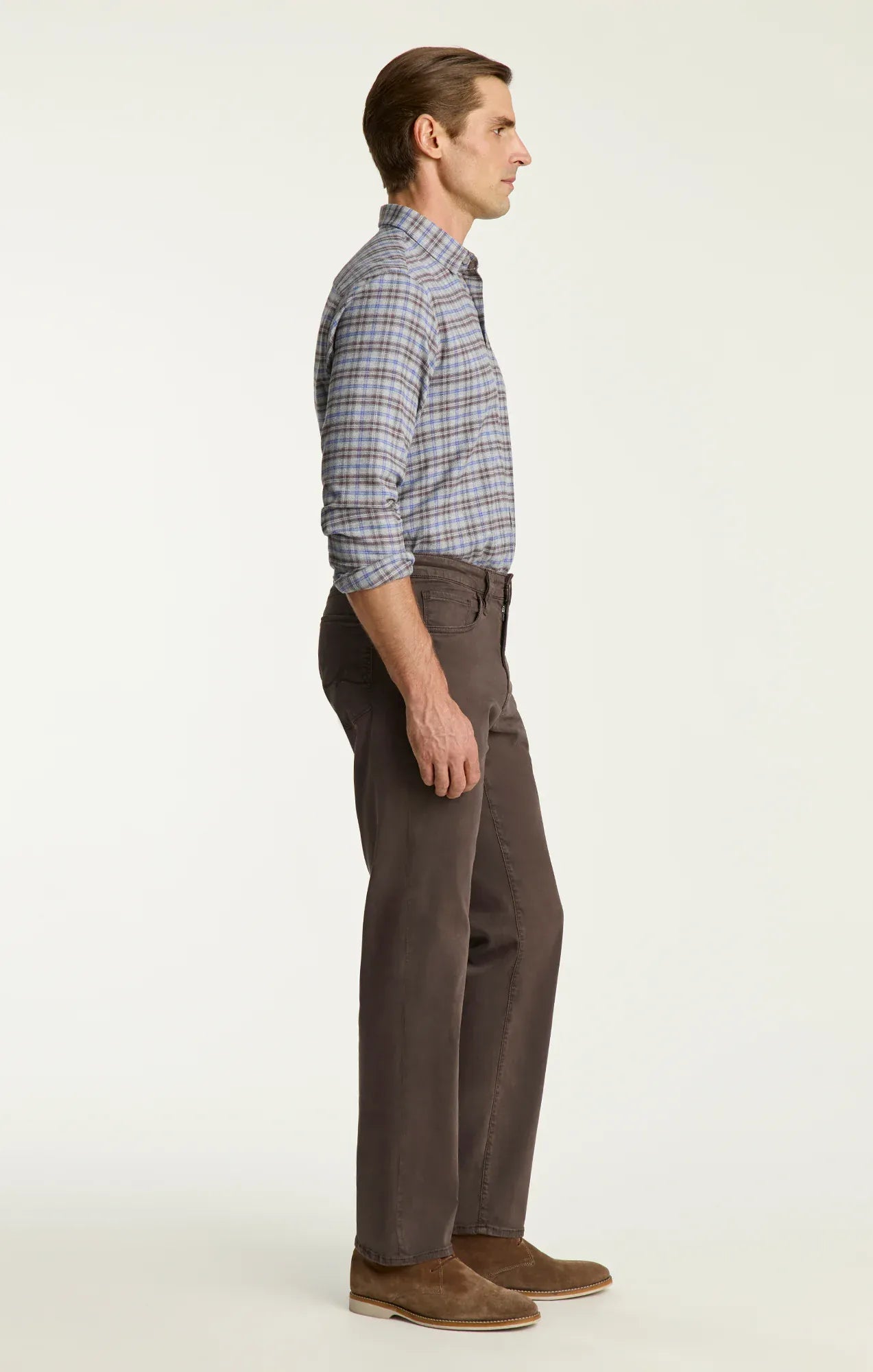 A man shown in profile wearing a blue and gray plaid button-down shirt, dark brown trousers, and brown suede shoes, standing against a plain off-white studio background.