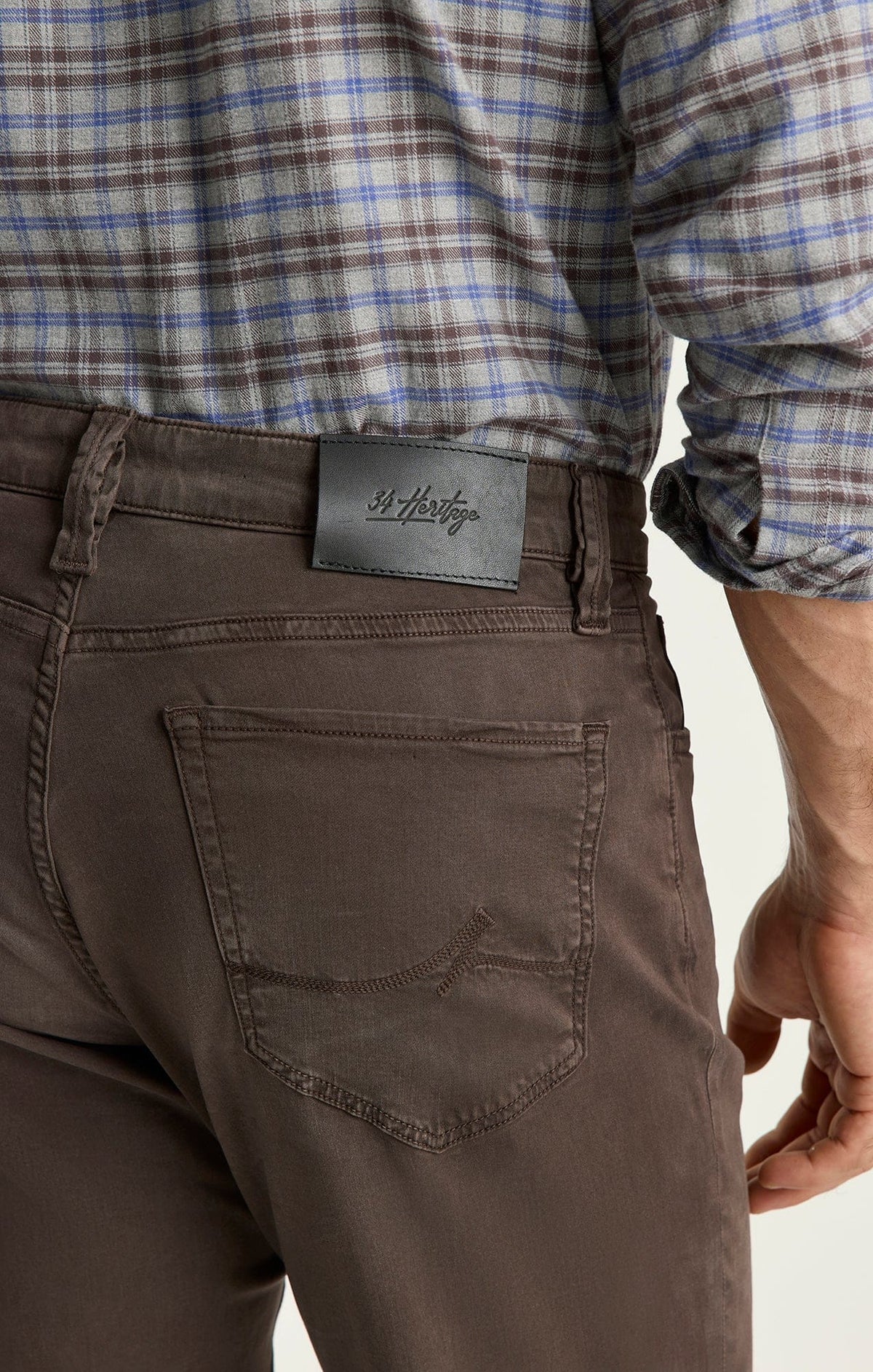 Close-up rear view of a person wearing dark brown chino pants with a black leather waistband patch featuring Chinese characters, paired with a blue and brown plaid shirt.