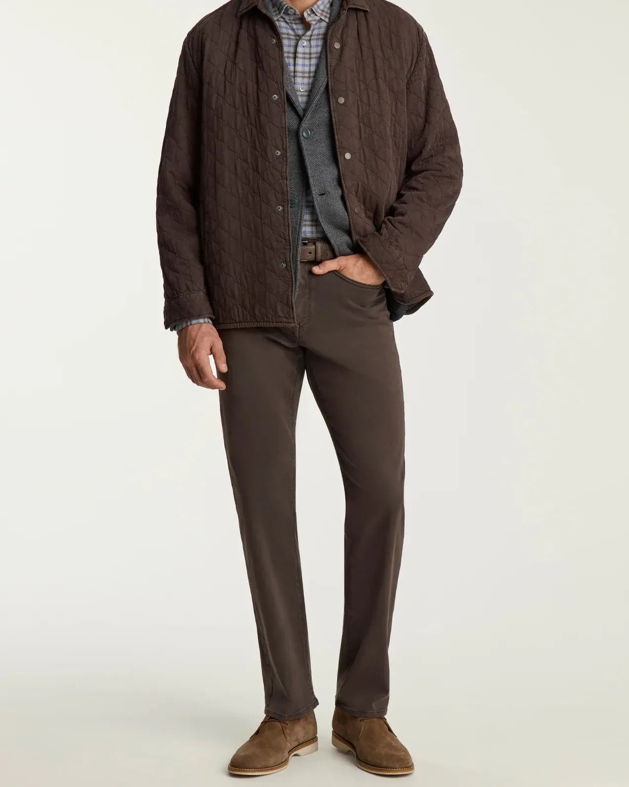 A man modeling a dark brown quilted overshirt jacket over a blue and white plaid shirt, gray vest, brown trousers, and brown suede shoes, photographed against a plain light gray studio background.