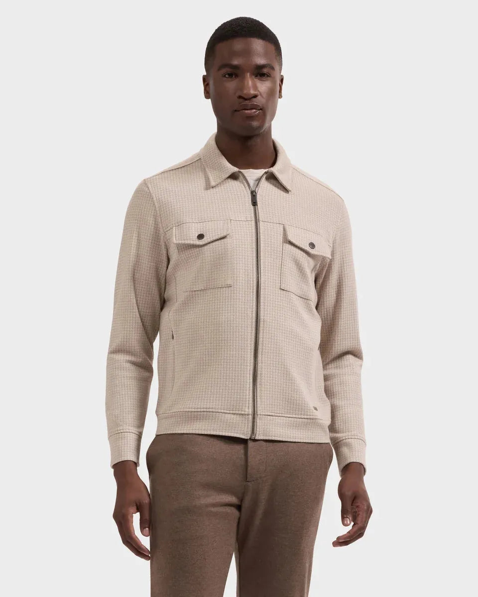 A man wearing a beige textured zip-up jacket with a collar and two chest pockets, paired with brown trousers, against a light gray studio background.