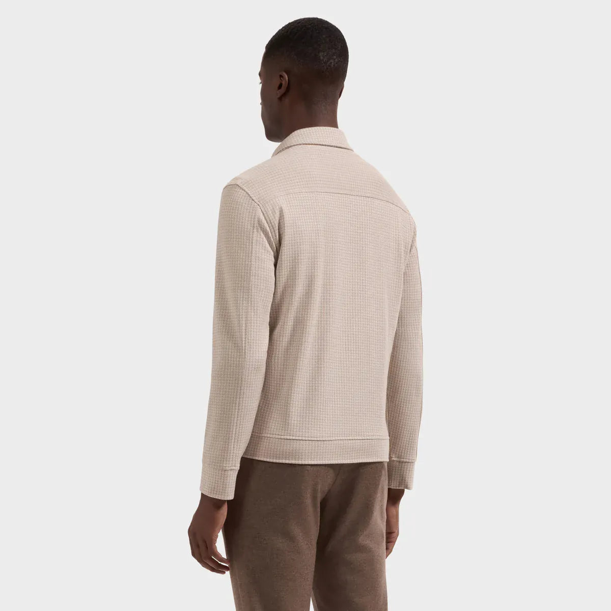 A man shown from the back wearing a light beige textured knit zip-up sweater and brown trousers, photographed against a clean light gray studio background.