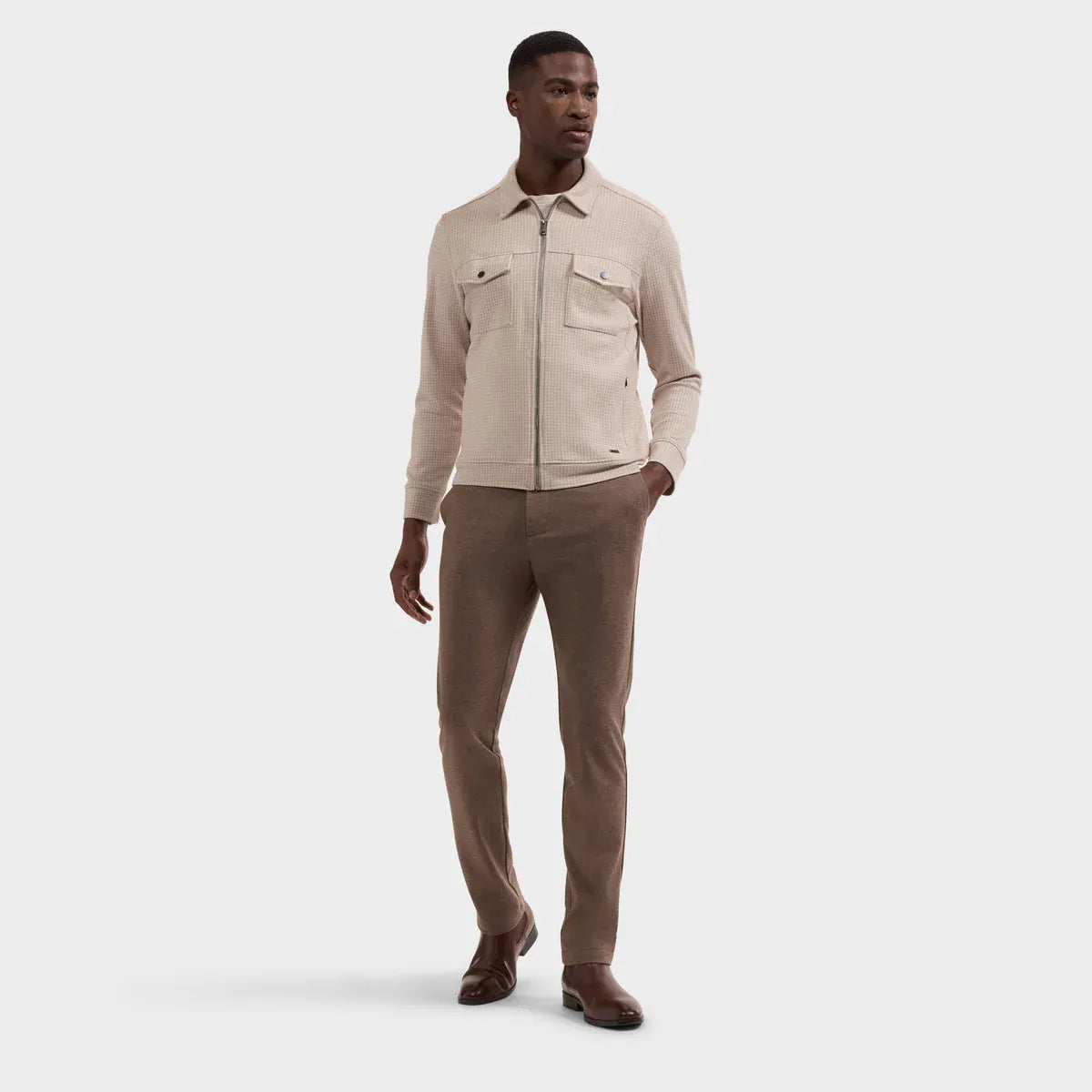 A male model wearing a light beige zip-up jacket and brown trousers, standing against a solid light gray background.