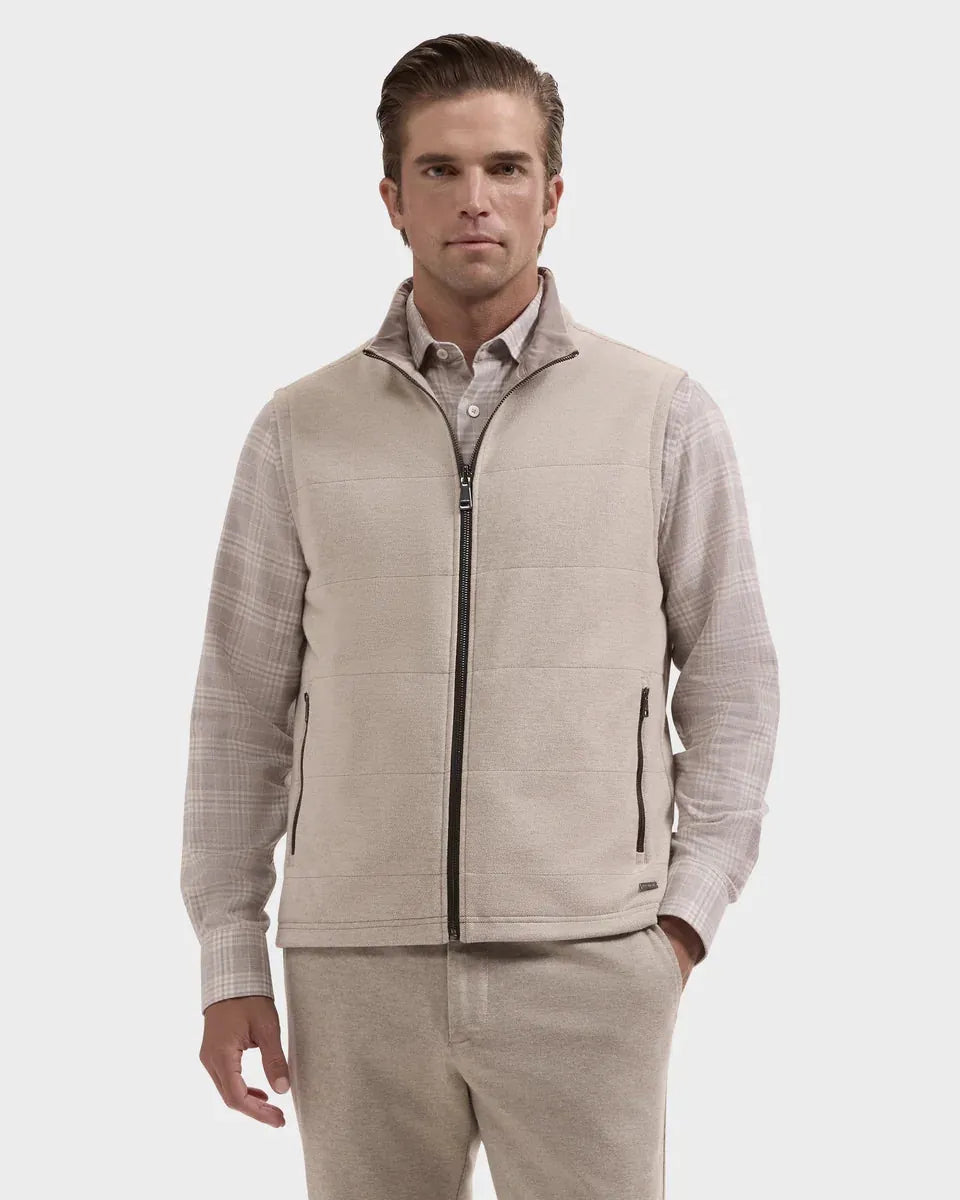 A man wearing a light beige zip-up vest over a beige plaid long-sleeve shirt and matching beige pants, photographed against a light gray studio background.