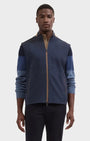 A man wearing a color-blocked zip-up fleece jacket in navy blue and medium blue with tan zipper accents, paired with dark pants, against a light gray studio background.