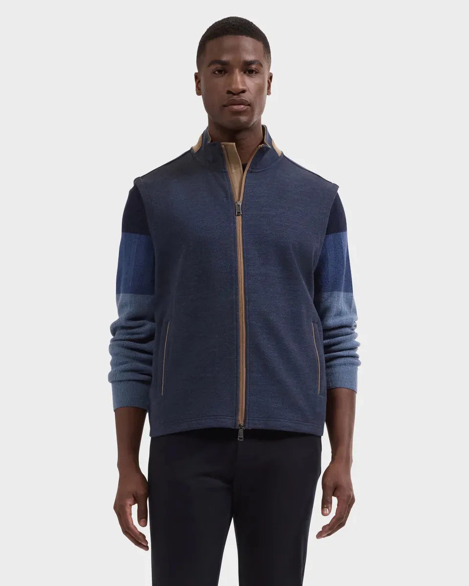 A man wearing a color-blocked zip-up fleece jacket in navy blue and medium blue with tan zipper accents, paired with dark pants, against a light gray studio background.