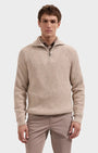 A man wearing a beige ribbed knit quarter-zip sweater with a stand collar, paired with light brown trousers, photographed against a solid light gray background.