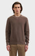 A man wearing a brown textured knit crewneck sweater and khaki pants, standing against a light gray studio background.