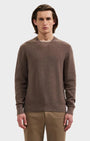 A man wearing a brown textured knit crewneck sweater and khaki pants, standing against a light gray studio background.