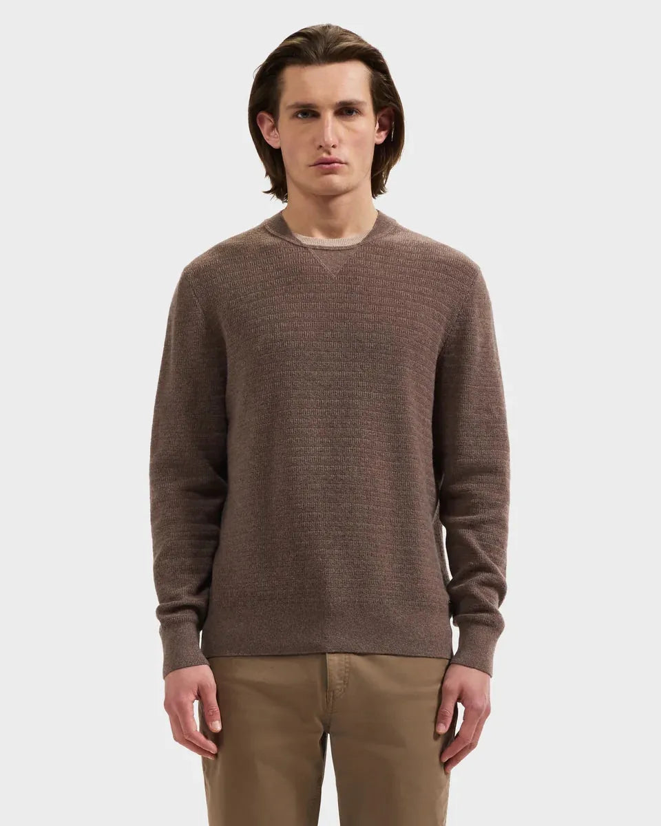 A man wearing a brown textured knit crewneck sweater and khaki pants, standing against a light gray studio background.
