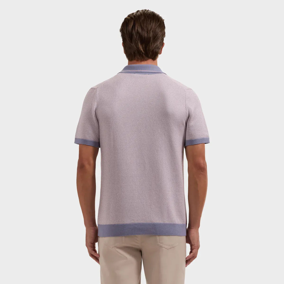A man viewed from the back wearing a light lavender knit polo shirt with contrasting dusty purple trim on the collar, sleeve cuffs, and hem, paired with beige trousers, against a solid light gray background.