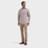 A man modeling a light beige long-sleeve button-down dress shirt with beige trousers and brown leather shoes, standing against a solid light gray background.